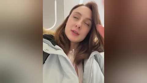 Video of a young woman with light skin and brown hair, wearing a light jacket, reclining in an airplane seat, eyes closed, appearing relaxed.