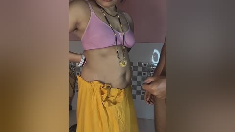 A video captures a young woman with light skin, wearing a pink bra and yellow skirt, adorned with multiple necklaces, standing in a dimly lit bathroom with a mosaic-tiled wall.