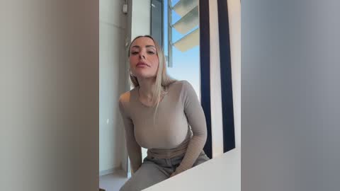 Video of a fair-skinned woman with long blonde hair, wearing a tight beige top, sitting in a modern, minimalist room with large windows and a blue sky visible outside.