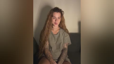 Video of a young woman with long, wavy brown hair, wearing a loose gray t-shirt, sitting on a dark couch, arms tattooed, making a playful face, indoors.