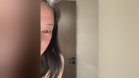 Video of a young Asian woman with long black hair partially visible, peering through a door frame, with beige tiled walls in the background.