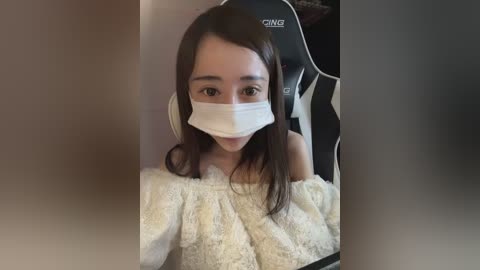 Video of an East Asian woman with long dark hair, wearing a white face mask, a white fuzzy sweater, and a black gaming chair.