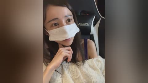 Video of an East Asian woman with straight, shoulder-length black hair, wearing a white surgical mask, beige cardigan, and a black gaming chair with an \"ACMG\" logo, sitting indoors.