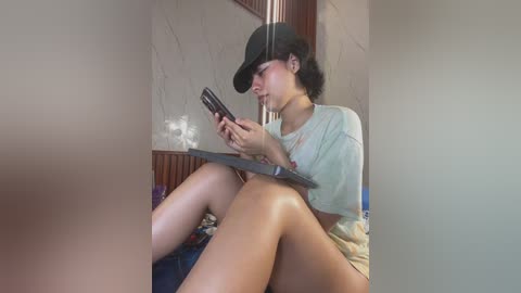Video of a young woman with curly hair, wearing a black hat and light-colored shorts, sitting on a bed, reading a book, with a wooden headboard in the background.