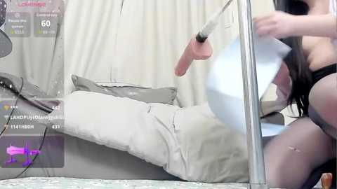 A digital image captures a nude woman with fair skin and long black hair, wearing only black thigh-high stockings, being restrained in a glass box. The background features white curtains and a gray bed.