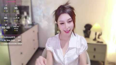 Video of a smiling East Asian woman with fair skin, wearing a white blouse, in a cozy room with a dresser, lamp, and a TV.