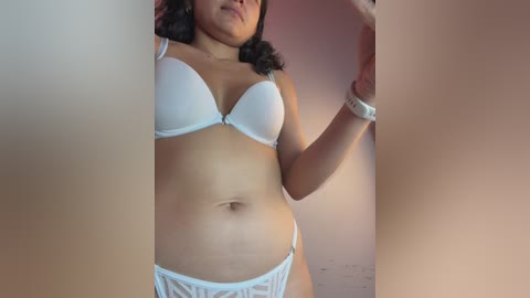 Video of a woman with medium skin tone, wearing a white bra and panties, standing in a dimly lit room with pink walls, partially obscured by a person's arm.