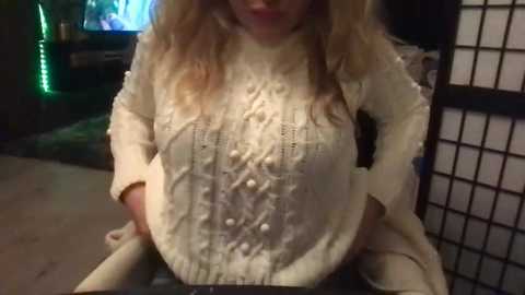 Video of a pregnant woman with long, wavy blonde hair, wearing a beige, knitted sweater, holding a baby in her arms. The background features a dimly lit room with a television displaying a colorful scene and a traditional Japanese shoji screen.