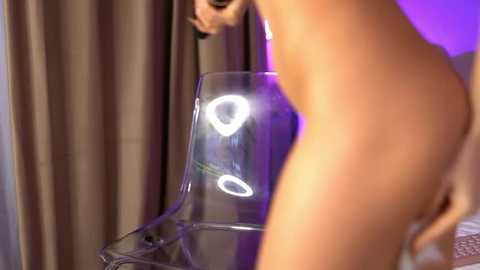 Media: A video of a nude, light-skinned person with a curvy, voluptuous figure, standing in front of a clear plastic chair with a circular light reflecting on its surface.