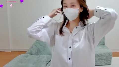 Video of an Asian woman with long brown hair, wearing a white button-down shirt, white mask, adjusting her mask, in a light-colored room with a sofa and a white wall.