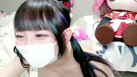 Video of an East Asian woman with long black hair, wearing a white surgical mask, sitting in a room with plush toys, including a pink doll with a red outfit.