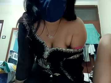 Media: Video of a woman with medium skin tone, black hair, wearing a blue mask, black off-shoulder dress exposing a pink bra, in a cluttered bedroom with a bed, clothes, and open door.