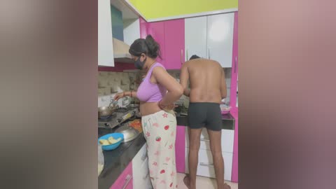 Video of a topless man in black shorts and a woman in a purple crop top and white pajama pants, cooking in a colorful kitchen with pink cabinets and a green ceiling.