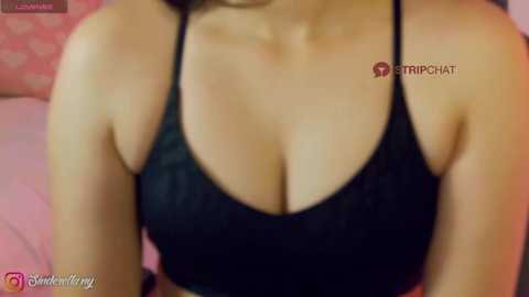 Video of a light-skinned woman with a medium build wearing a black sports bra, highlighting her ample breasts. The background is blurred, showing pink and white patterns.