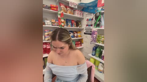 A candid video of a young woman with light skin and brown hair, wearing a light gray off-shoulder top, standing in a cluttered convenience store, surrounded by shelves stocked with various products.