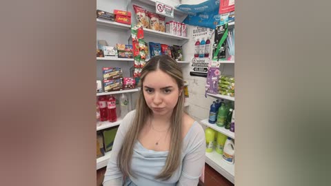A video of a young woman with long blonde hair, wearing a light blue top, sitting in a cluttered, colorful toy store with shelves filled with various toys, games, and snacks.