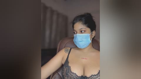A video of a woman with medium brown skin, wearing a blue face mask and black lace bra, seated in a brown leather chair.