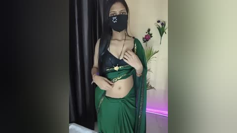 Video of a young woman with medium skin tone, dark hair, wearing a green sari with a black lace bra, black face mask, and green shawl, standing in a dimly lit room with a black curtain, flowers, and pink lighting.
