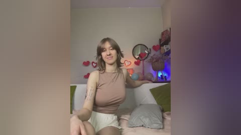 Video of a young woman with medium-length brown hair, light skin, and a slim build, wearing a beige tank top and white shorts, sitting on a bed adorned with heart decorations and a green pillow.