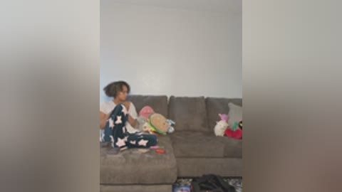 Video of a young Black girl with short hair in blue pajamas, sitting on a beige sectional sofa in a cluttered living room.