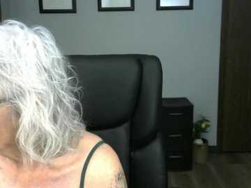 Video of a woman with shoulder-length, white, curly hair, wearing a black strapless top, sitting in a black leather chair, with a beige cabinet and framed artwork on a light grey wall in the background.