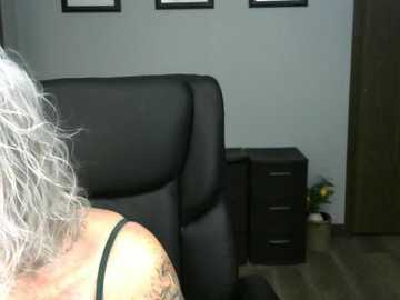 Video of a gray-haired woman in a black bra, sitting on a black leather office chair, next to a dark wooden file cabinet in a minimalist office with framed pictures on a light gray wall.