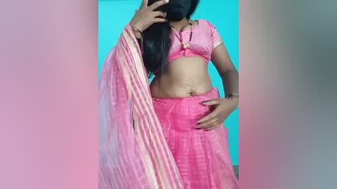 Video of a South Asian woman with medium brown skin, wearing a pink and white striped sari, standing in front of a turquoise wall, hands on hips.