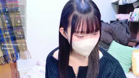 Video of a young Asian woman with long black hair, wearing a face mask, sitting indoors. She's dressed in a dark sweater. Background shows a plaid blanket, books, and a bed.