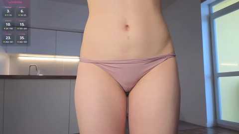 Video of a woman's bare midriff and lower torso in light pink panties, standing in a modern kitchen with white cabinets and a window.