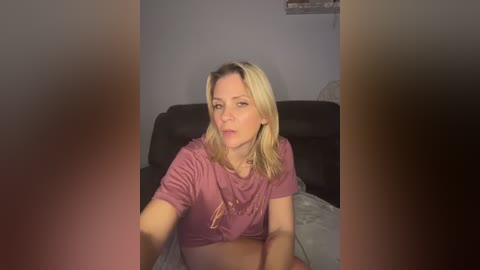 A video of a blonde woman with shoulder-length hair, wearing a pink T-shirt, seated on a black couch in a dimly lit room.