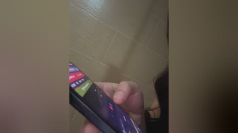 Video of a person holding a smartphone with a colorful game screen, against a beige tiled wall, with a blurred, dark-haired person partially visible in the background.