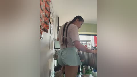Video of a young woman with long braids in a kitchen, wearing a white t-shirt and light blue shorts, washing dishes in a cluttered room with a green wall and red posters.