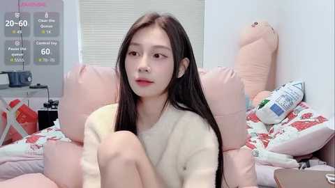 Video of a young Asian woman with long black hair, wearing a cream sweater, sitting on a pink and white patterned bed, surrounded by stuffed animals and a digital clock.