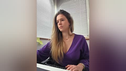 Video of a Caucasian woman with long, light brown hair, wearing a purple V-neck blouse, playing a keyboard in a bright office with white blinds and yellow sticky notes on the wall.