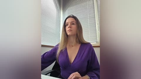 Video of a light-skinned woman with long blonde hair, wearing a deep purple, low-cut blouse, sitting in front of closed blinds, looking serious.