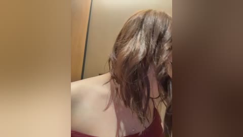 Video of a woman with shoulder-length brown hair, wearing a red off-shoulder top, looking down, in a dimly lit, beige-walled room.