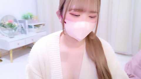 Video of a young Asian woman with long brown hair, wearing a white mask and a light sweater, sitting in a softly lit, white room with a vase of flowers on a table.