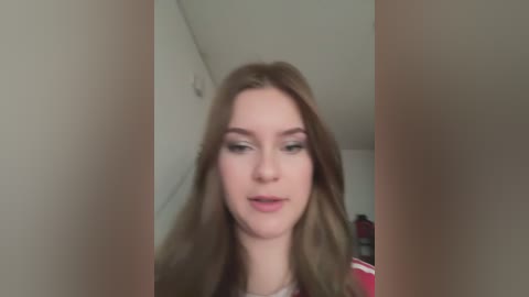 A video of a young woman with long, straight brown hair, light skin, and a neutral expression. She's wearing a red top and is standing in a dimly lit, indoor setting with white walls.