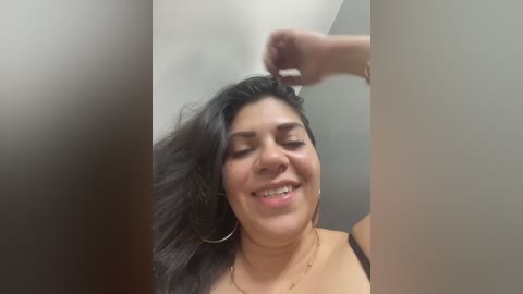 A video of a smiling Latina woman with long dark hair, wearing hoop earrings and a black bra, in a brightly lit room.