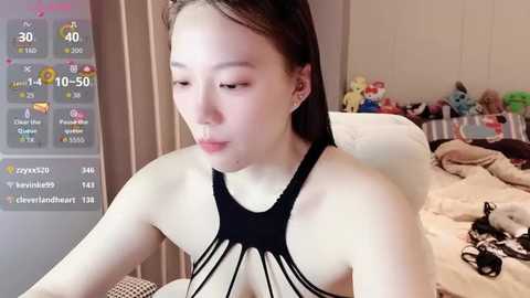 A video of a young Asian woman with light skin and straight brown hair, wearing a black crochet halter top, sitting on a bed with a messy background.