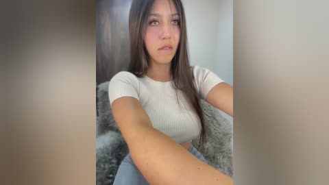 Video of a young woman with long brown hair, wearing a tight, ribbed white crop top and high-waisted jeans, sitting on a gray, fluffy rug in a dimly lit room.