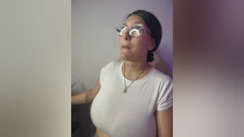Video of a woman with medium-sized breasts, wearing a white top and glasses, standing against a blurred background, possibly indoors.