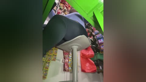 Video of a person in black leggings and a blue shirt sitting on a white stool, surrounded by brightly colored candy wrappers and a green tarp, in a cluttered indoor space.