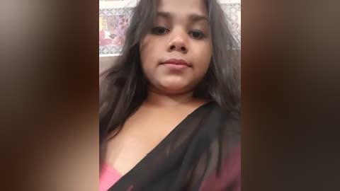 Video of a young South Asian woman with medium-brown skin, long black hair, and a subtle smile, wearing a black sari with a pink blouse, set against a tiled bathroom background.