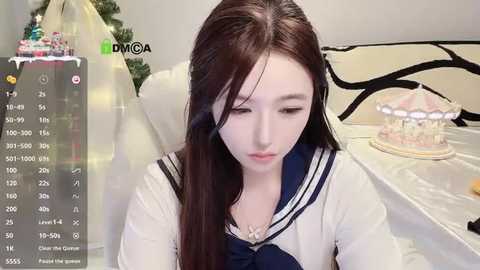 Video of a young Asian woman with long black hair and fair skin, wearing a white sailor-style school uniform, sitting on a white chair. Background features a decorated Christmas tree and a cake on a table.