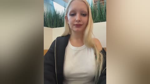 Video of a young, fair-skinned woman with long, straight blonde hair, wearing a white T-shirt and black jacket, standing in front of a green potted plant and beige wall.