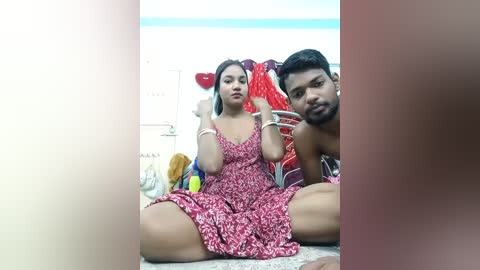 Video of a young couple, a woman in a floral dress and a man shirtless, sitting on the floor in a dimly lit room with colorful textiles behind them.