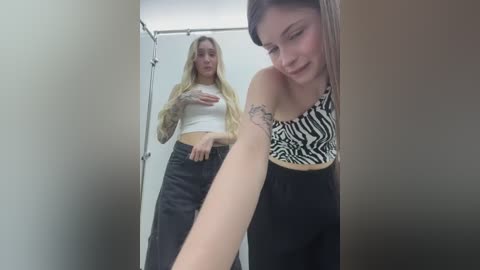 Media: Video of two young women in a changing room: one with long blonde hair, a white crop top, and black pants; the other with straight, brown hair, a zebra-print top, and black pants, looking at her arm.