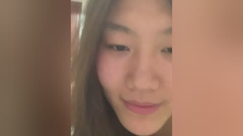 A close-up video of a young Asian woman with fair skin, long brown hair, and light makeup, smiling slightly. The background is blurred, with beige tones dominating. The image is slightly out of focus.