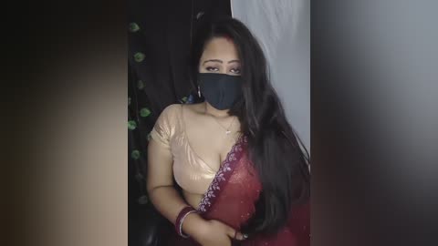 Video of a South Asian woman with medium skin tone and long black hair, wearing a gold and red saree, black mask, and silver earrings, indoors against dark backdrop.